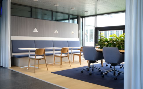 Eine Fotografie aus der umgestaltetetn Büronische mit Sitzbank, Tischen, Teppich und Pflanzentisch. 